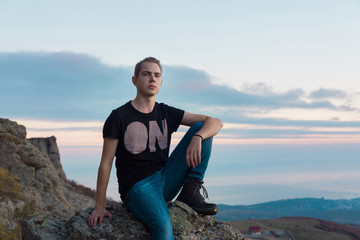 Naklejka na ściany i meble Young attractive man located with on the top of mountains against the backdrop against a blue and pink sunset, looking towards the camera