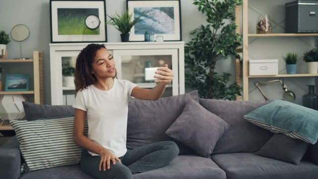 Young African American lady is calling friends on skype making online video call with smartphone talking and showing her new apartment with cool interior.