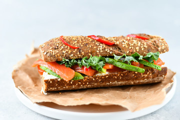 sandwich from a cereal baguette with avocado, salmon, cream cheese, tomatoes and lettuce leaves on a white plate.  light background, selective focus and copy space.