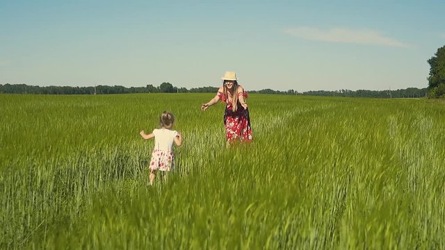 Slow motion: Young mother plays with a child in the field, dressed in a red dress that flutters in the wind. The daughter goes to her on the green grass, the mother pulls her hands, smiles.