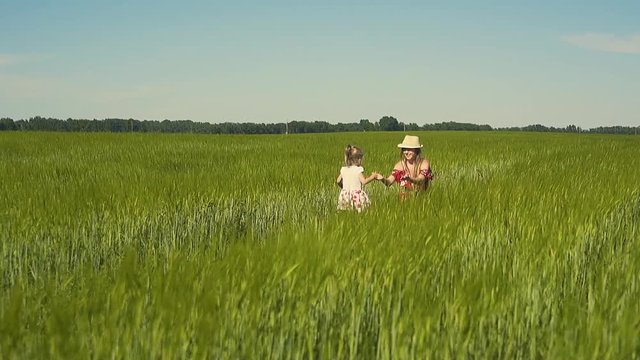 Slow motion: Young mother plays with child in the field, dressed in red dress. The daughter goes to her on green grass, the mother pulls her hands, smiles. Then she lifts her in arms and turns.