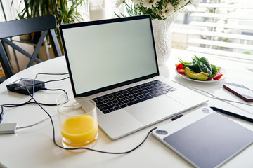 Opened laptop, graphics tablet and breakfast with juice on white table, empty screen for mockup design.