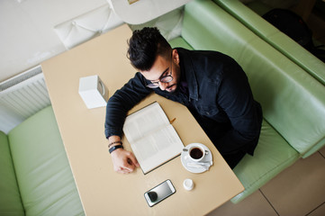Arab man wear on black jeans jacket and eyeglasses sitting in cafe, read book and drink coffee....