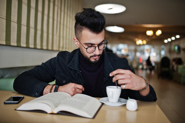 Arab man wear on black jeans jacket and eyeglasses sitting in cafe, read book and drink coffee. Stylish and fashionable arabian model guy.