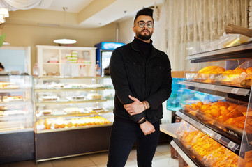 Arab man wear on black jeans jacket and eyeglasses in cafe choose bakery. Stylish and fashionable...
