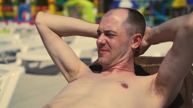 Close-up, Portrait: Bald Italian man is lying on the beach, on a sun lounger, sunbathing. Frowning from the sun, looking away.
