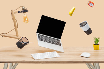 laptop with blank screen and stationery levitating in air at workplace isolated on beige