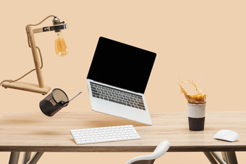 laptop with blank screen and lamp levitating in air above workplace with thermomug with coffee splash isolated on beige
