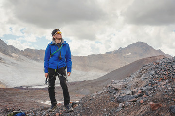 Obraz premium Portrait of a professional guide of a mountaineer in a cap and sunglasses with an ice ax in his hand against the background of the sky and the snow-capped mountains of the Caucasus