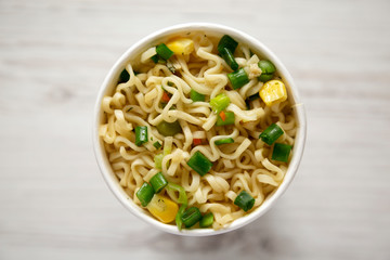 A paper cup of instant ramen noodles over white wooden surface. Flat lay, from above, overhead. Close-up.