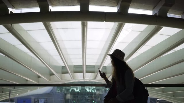 Side view of young woman silhouette in hat browsing on mobile phone in sun shine from windows. Concept modern train station or terminal