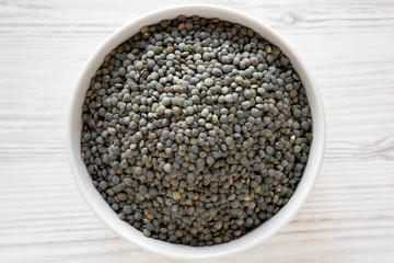 Dry green french lentils in gray bowl over white wooden background, top view. Flat lay, overhead, from above. Close-up.