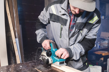 A young carpenter with a beard works with an electric plane without suctioning sawdust. Leveling and sanding wooden beams