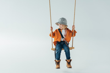 cute kid in jeans and orange shirt sitting on swing and looking away on grey background - obrazy, fototapety, plakaty