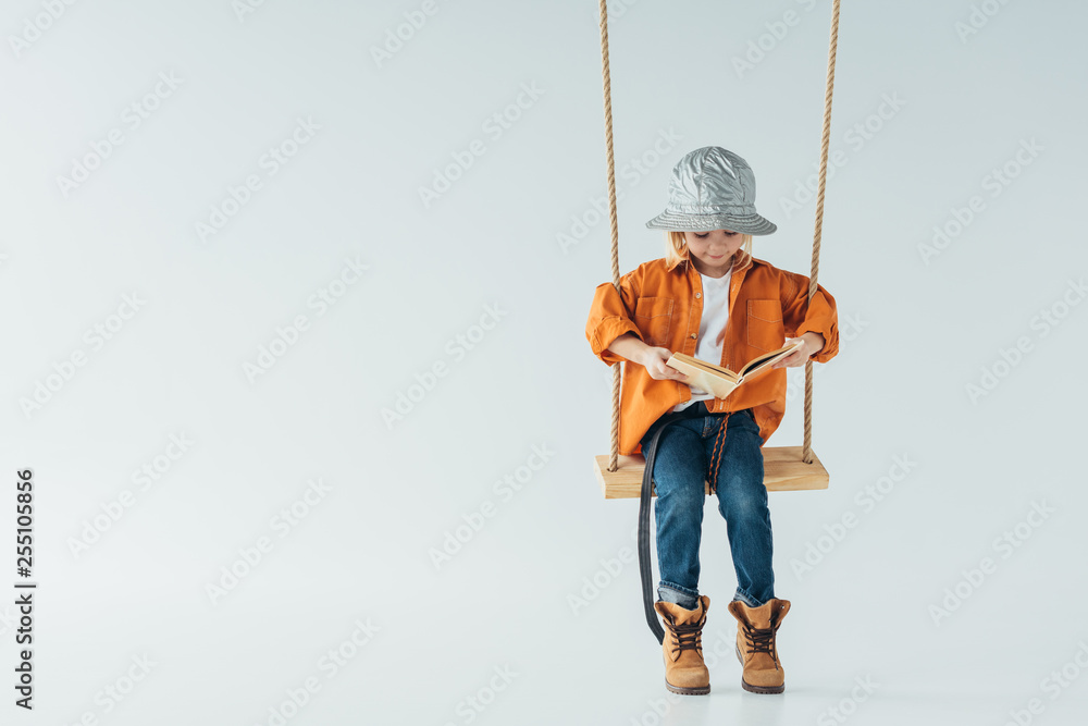 Wall mural cute kid in jeans and orange shirt sitting on swing and reading book on grey background