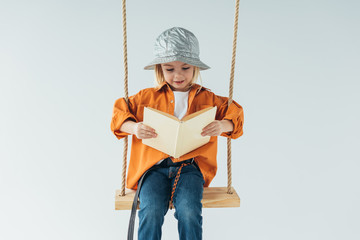 adorable kid in jeans and orange shirt sitting on swing and reading book isolated on grey