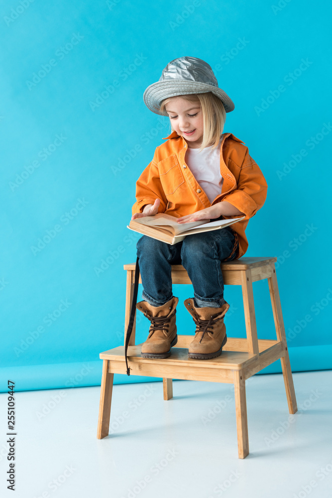 Wall mural kid in jeans and orange shirt sitting on stairs and reading book on blue background - Wall murals