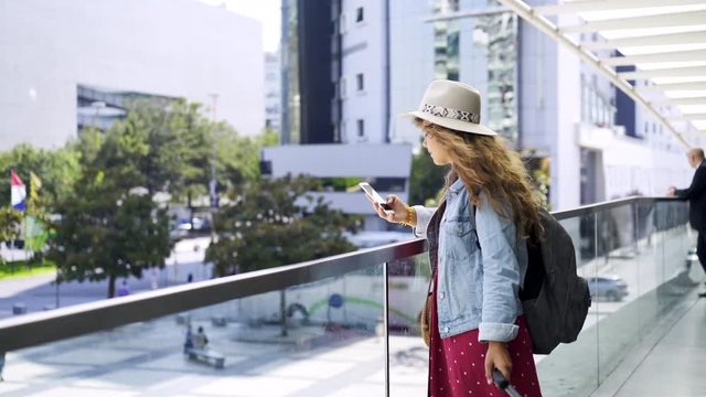 Crop view of young woman in hat with luggage browsing on mobile phone near open window in sunny day inside interior of modern transport terminal