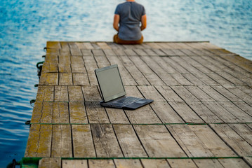 Young man sitting on a pier, using his laptop. Work and vacation.