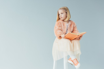  kid in faux fur coat and skirt sitting on highchair and holding book isolated on grey