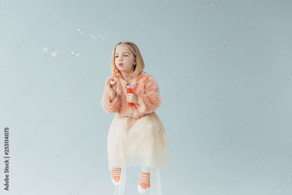 Wall mural adorable kid in faux fur coat and skirt sitting on highchair and blowing soap bubbles isolated on grey