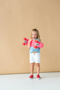 Smiling And Cute Kid In Shirt And Shorts Holding Pink Penny Board On Beige Background