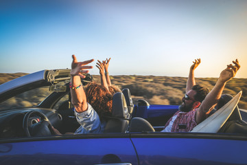 Side view group three euphoric friends in convertible car twisting and waving Two curly girls and charming bearded man on vacation having fun in topless auto Arms up palms open driving in the sunlight - obrazy, fototapety, plakaty