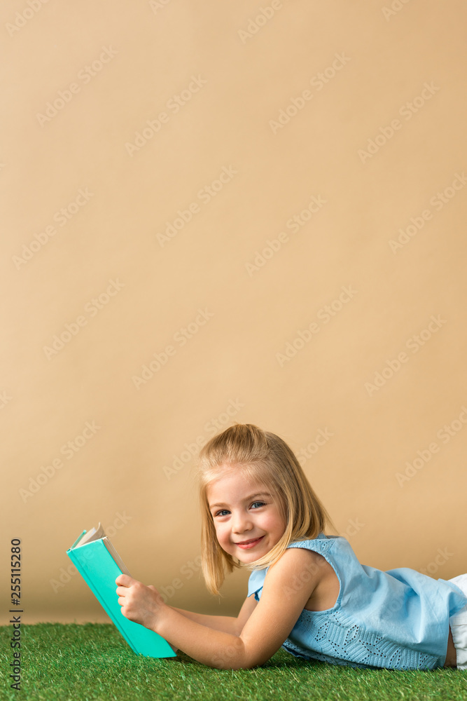 Wall mural smiling and cute kid lying on grass rug and holding book on beige background