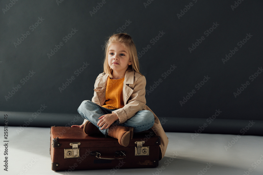 Wall mural pensive child sitting with crossed legs on leather suitcase on black background