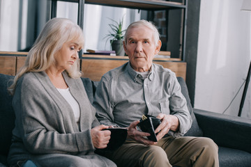 upset senior couple sitting on couch and holding wallets at home