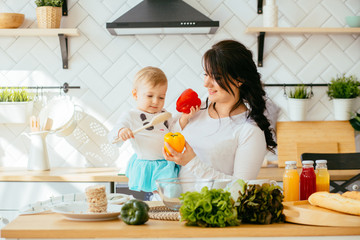 Kid and mother cooking together. Mother holding her little toddler girl and playing in a white kitchen with modern oven. They ares making salad with peppers for dinner.