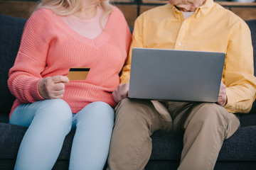 cropped view of senior couple sitting on couch with laptop and credit card while doing online shopping at home