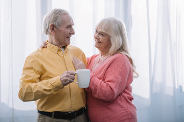 smiling senior woman with grey hair looking at camera and embracing man with cup of tea at home
