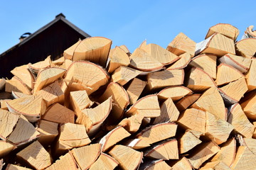 A pile of birch wood and the roof of the village house. Fresh and bright woodpile of birch fire wood. In the background, the old roof of the house. Blue sky. Close up.