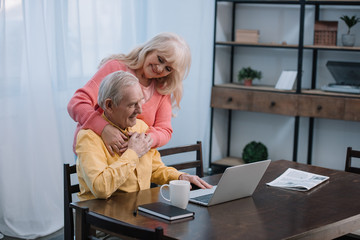 happy senior couple hugging while man sitting at table and using laptop in living room