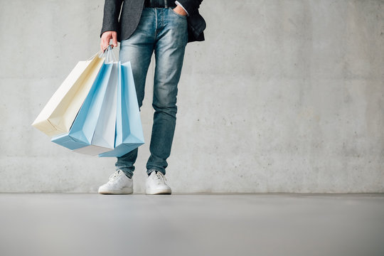 Fashion Shopping Lifestyle. Man Legs In Jeans Cropped Shot. Paper Bags Assortment. Copy Space On Grey Background.