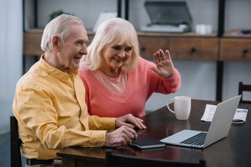 smiling senior couple sitting at table and using laptop during video call at home