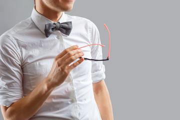 Stylish and fashionable man in a white shirt with a bow tie holding glasses in his hand. Elegant man posing in studio standing against gray background. Men's classic eyewear fashion. Gesticulation.