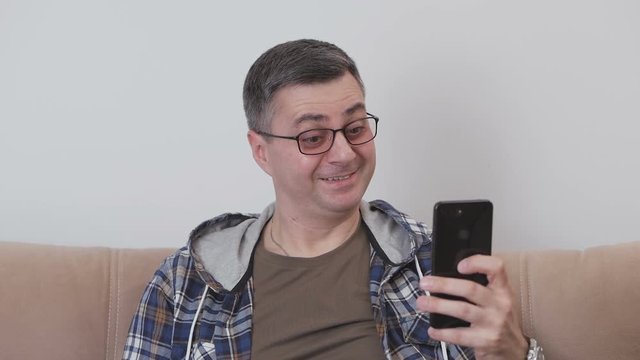 A middle-aged man, sitting on a couch, takes a picture of himself using a smartphone, makes faces and makes a stupid face.