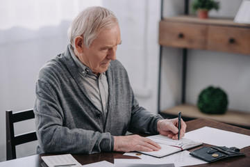 senior man sitting at table with paperwork and writing in notebook