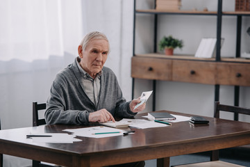 senior man sitting at table with paperwork and using calculator while counting money at home