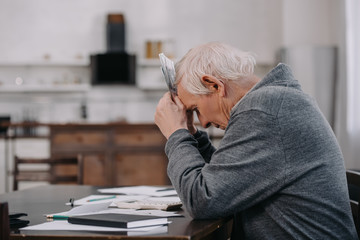 stressed senior man in casual clothes sitting at table with paperwork and holding money at home - obrazy, fototapety, plakaty