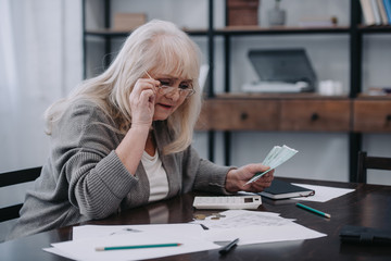 senior woman sitting at table with paperwork, holding glasses and counting money