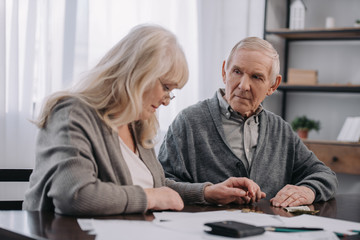 Senior couple in casual clothes sitting at table with paperwork while counting money