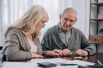 Senior couple in casual clothes sitting at table with paperwork while counting money