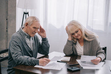 stressed senior couple with hands on head sitting at table and looking at bills