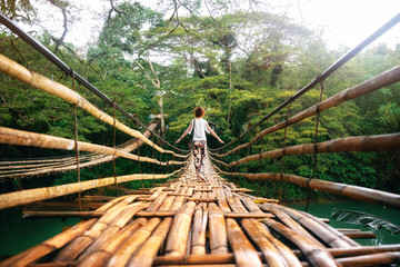 Back view of young woman on suspension wooden bamboo bridge across Loboc river in jungle. Vacation...