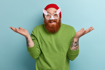 Funny cheerful bearded man wears round rabbit glasses with ears, has tattoos, spreads palms, doesnt know what to do, wears red headgear and green sweater, isolated over blue wall, has fun indoor