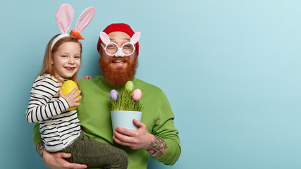 Family celebration concept. Satisfied funny bearded man in white bunny spectacles holds vase with green plant and dyed eggs, carries little girl with rabbit ears, have annual Easter egg hunt