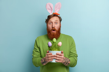 Stupefied bearded adult man wears bunny long ears on head, carries Easter symbols, opens mouth from...